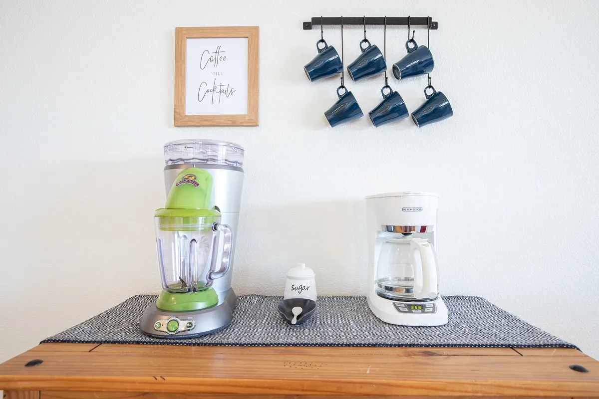 A bright and welcoming kitchen nook featuring a coffee maker, blender, sugar container, and hanging mugs, with framed artwork adding a personal touch. The compact area is thoughtfully organized for ease and convenience, highlighting the stocked essentials and inviting ambiance.