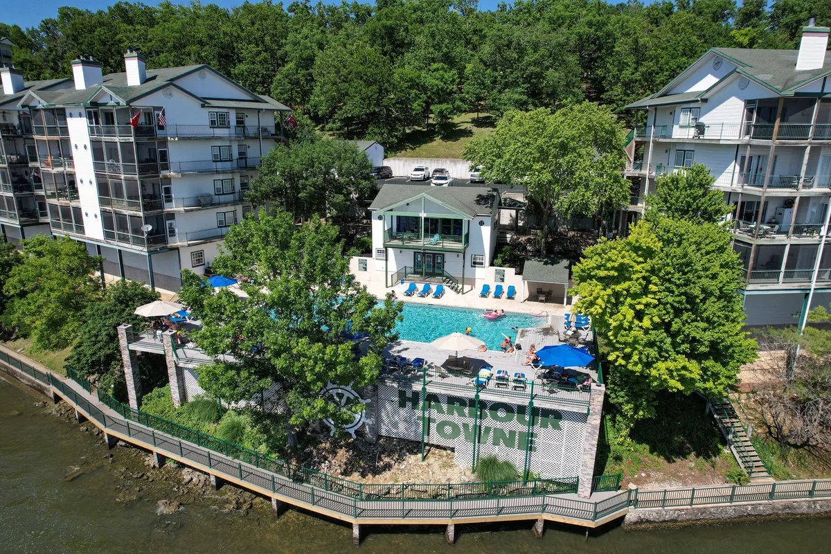 A vibrant exterior scene featuring the lake-view pool at Harbour Towne. The pool is surrounded by lounge chairs and shaded by tall trees, with the Harbour Towne sign visible. Guests can be seen swimming, sunbathing, and socializing, all set against a picturesque lakeside backdrop.