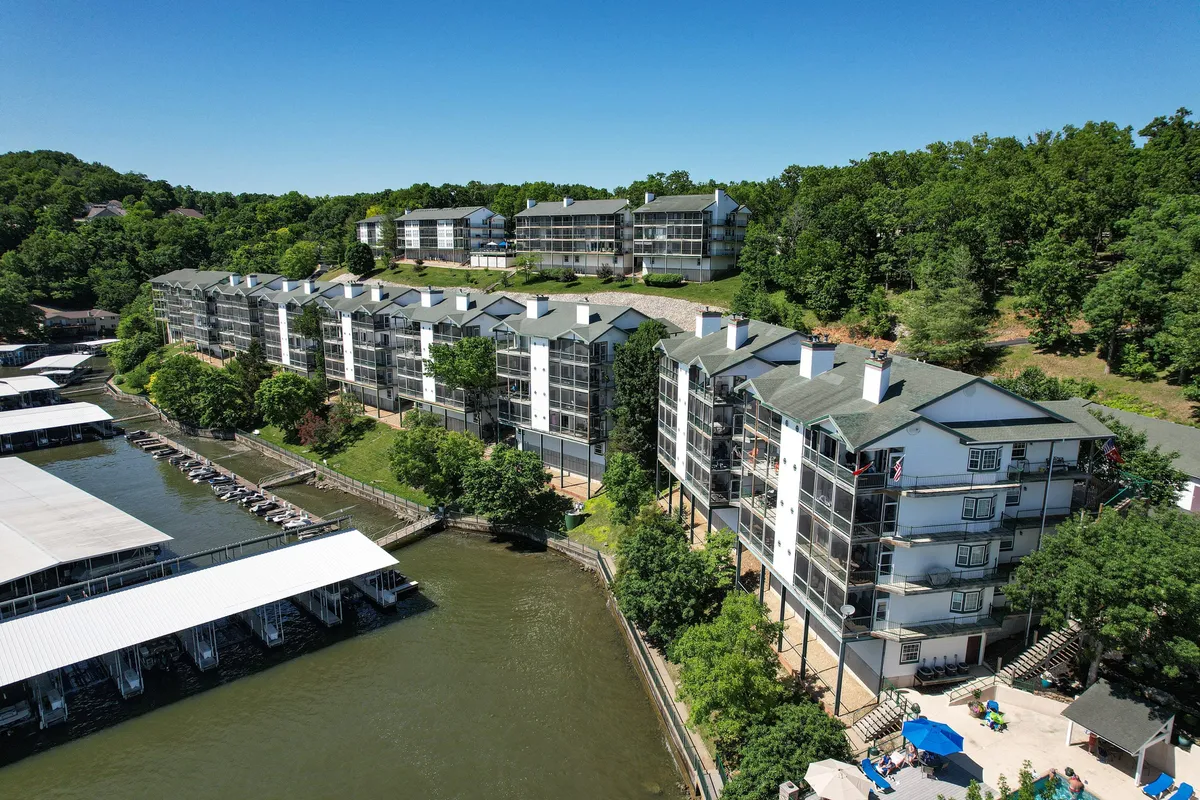 A panoramic exterior view showing lakefront buildings, the marina, and a lush green hillside. The scene captures the proximity to water activities and the natural beauty surrounding the property, with areas for boating, fishing, and swimming nearby.