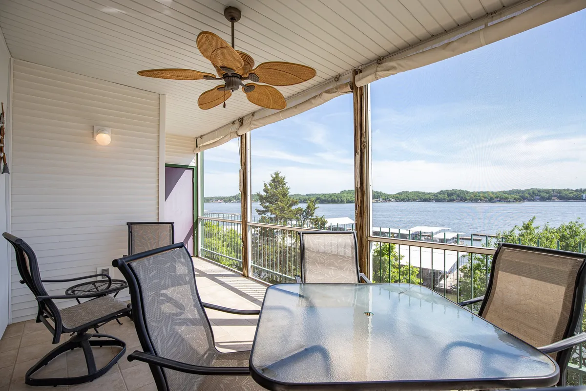 An exterior shot of a lakefront balcony with outdoor seating and a glass table, offering a panoramic view of the water. The space is set up for both dining and relaxation, with a ceiling fan for comfort and the lake serving as a dramatic backdrop.