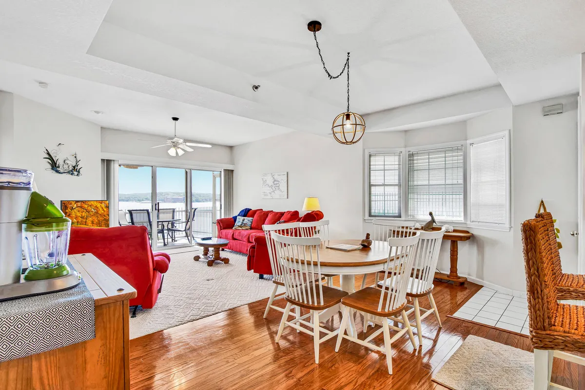 This image displays a cozy living room with a red sofa and a dining table set beside large windows that overlook the lake. The expansive windows invite abundant natural light and frame a beautiful water view, while the open-plan layout seamlessly connects the living and dining spaces.