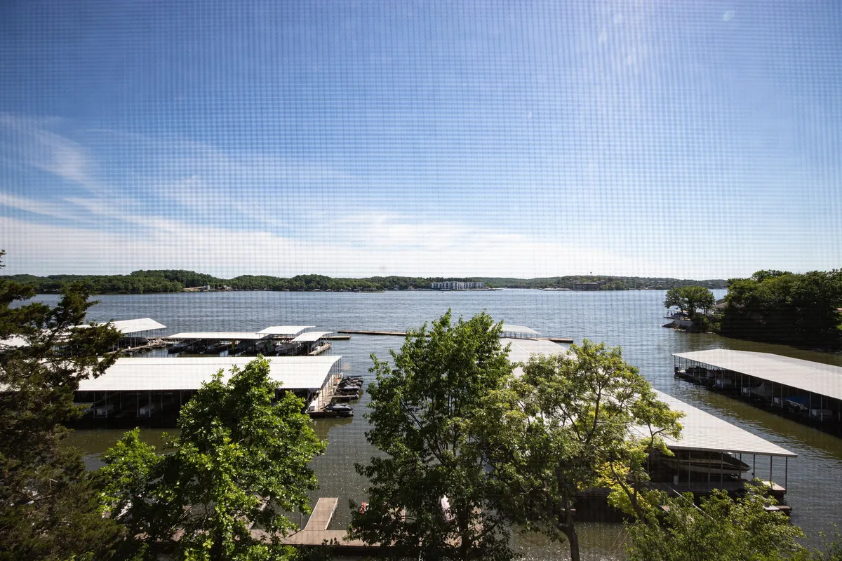 Expansive view of the lake and marina, bordered by green trees, capturing the tranquil beauty and recreational opportunities of the Lake of the Ozarks. The scene encourages boating, morning coffee moments, and sunset reflections.