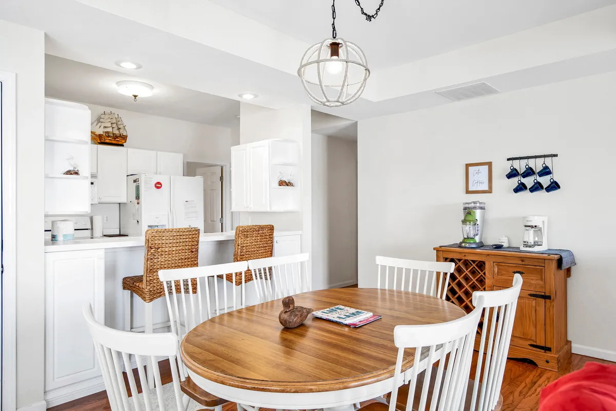 A dining area with a round wooden table, white dining chairs, basket weave bar stools, and a wooden sideboard. The arrangement hints at group gatherings and celebrations, providing a sense of the welcoming, social atmosphere described in the section.