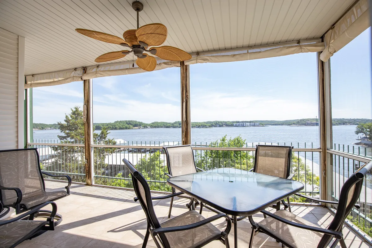 The image shows an inviting balcony overlooking the sparkling waters of Lake Ozark, furnished with a table and comfortable chairs beneath a ceiling fan—perfect for outdoor dining or relaxing. The expansive view captures the serene main channel, ideal for watching sunrises or sunsets. The setting exudes tranquility and encourages moments of connection with family and friends.