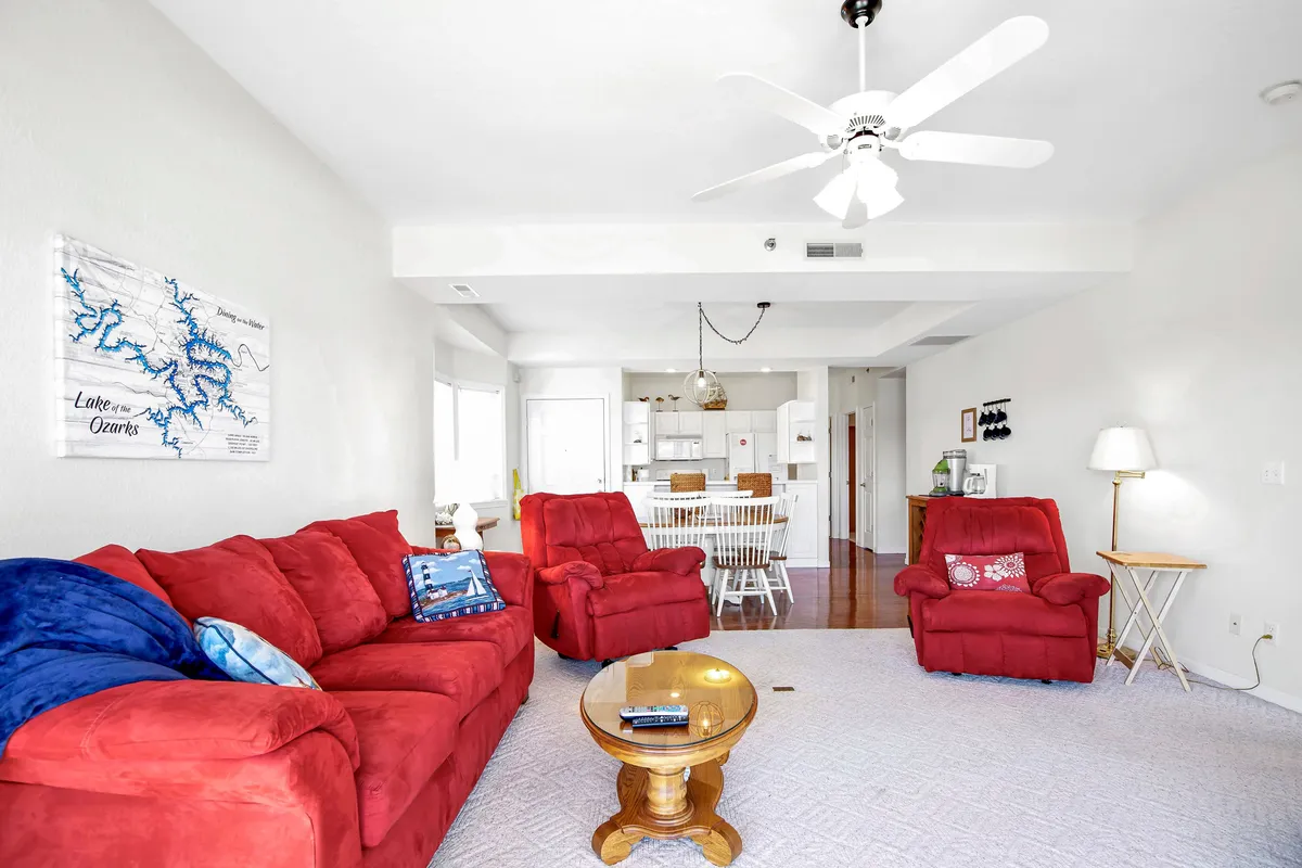 A warm and welcoming living area is shown with red sofas, a wooden coffee table, and a kitchen area visible in the background. Artwork on the wall adds a personal touch, and the open layout encourages relaxation and conversation.