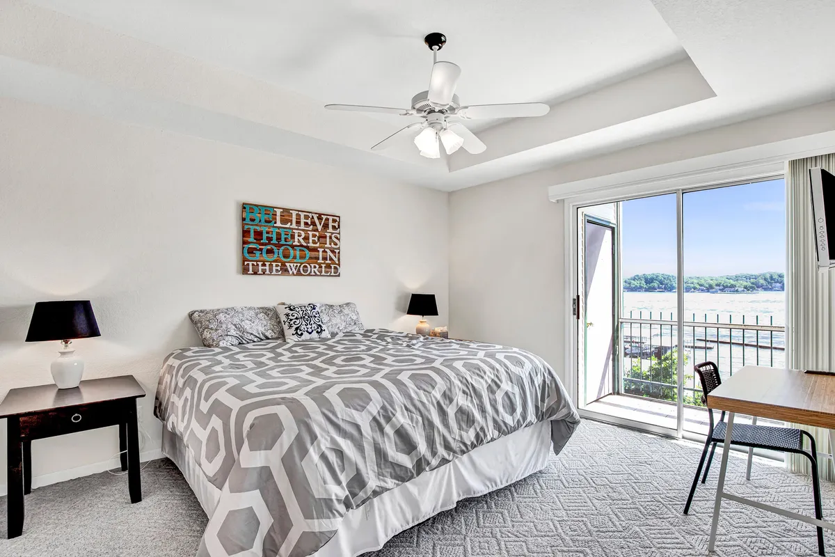 A plush bed with a patterned duvet sits beneath a large window offering beautiful outdoor views, with motivational wall art adding a personal touch. The scene captures a serene, airy atmosphere ideal for restful sleep and quiet reflection, while the window hints at the lake view and balcony access described in the section.